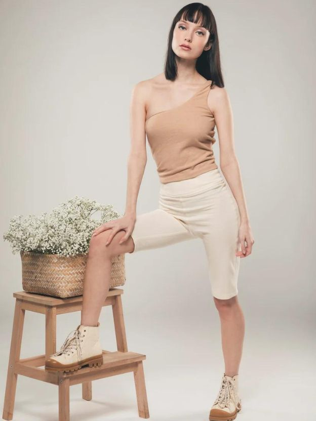 Woman standing leg on a stool with white flowers, woman wearing beige one shoulder top and white shorts from Organique