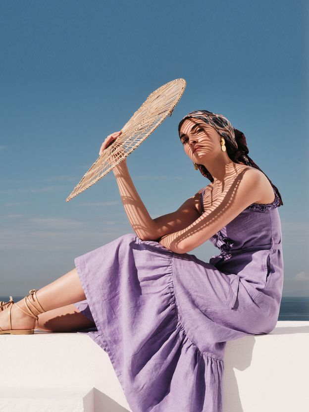 Nainen poseeraa ulkona laventelin värisessä pellavamekossa viuhka kädessä