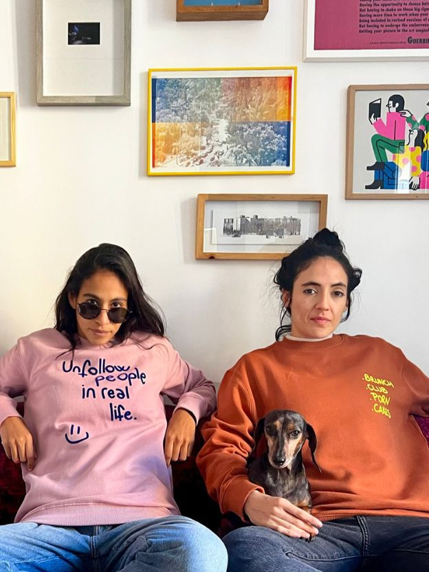 Two women sitting on a couch with a dog wearing Raiz shirts