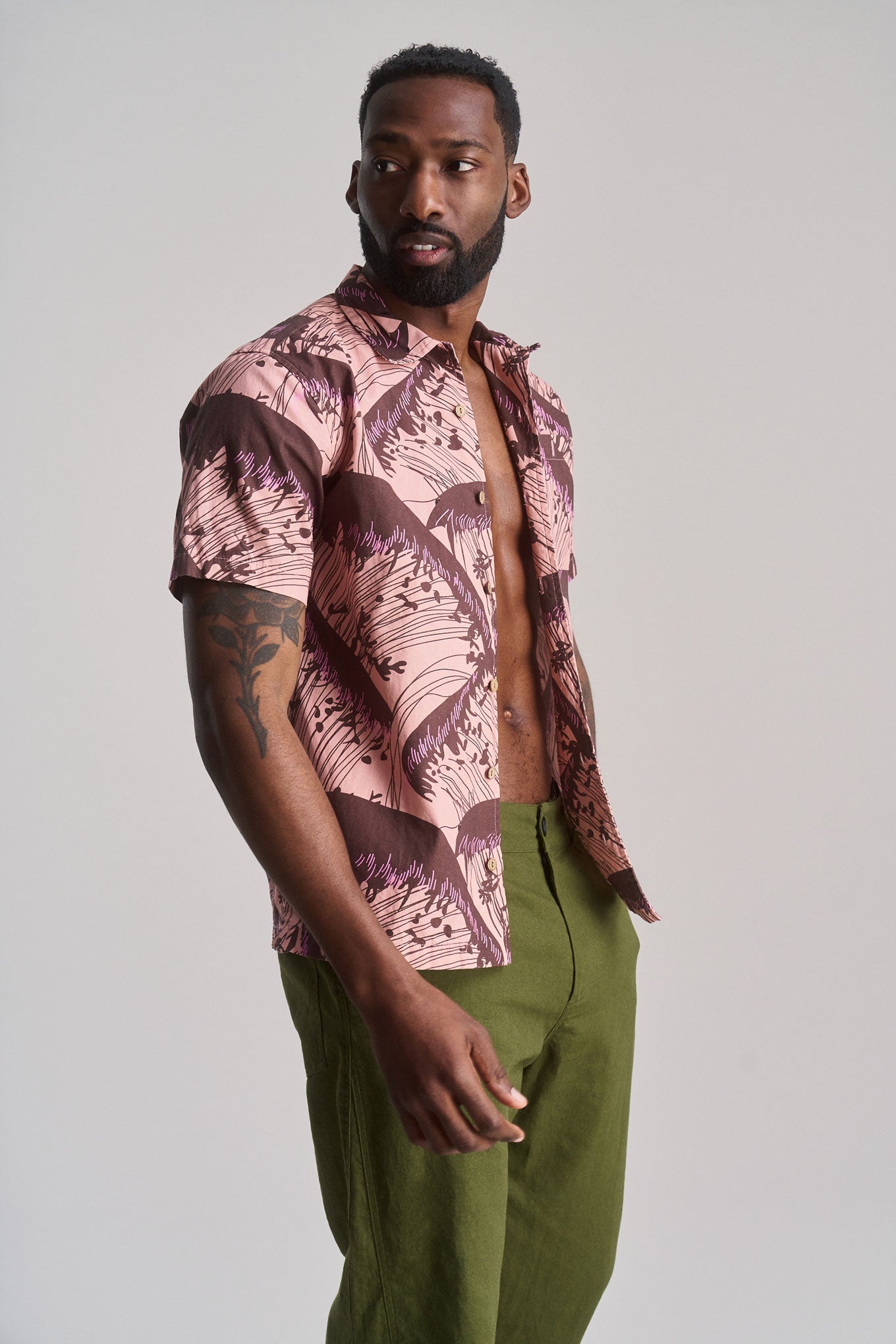 A bearded man with a tattoo models the SPINDRIFT Organic Cotton Shirt in Pink Wave and olive green pants against a plain light gray background.