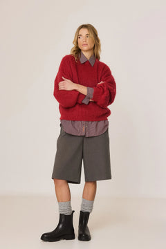 A model stands against a plain light beige studio background wearing a chunky red knit sweater over a gray collared shirt, paired with gray shorts, gray socks, and black boots.