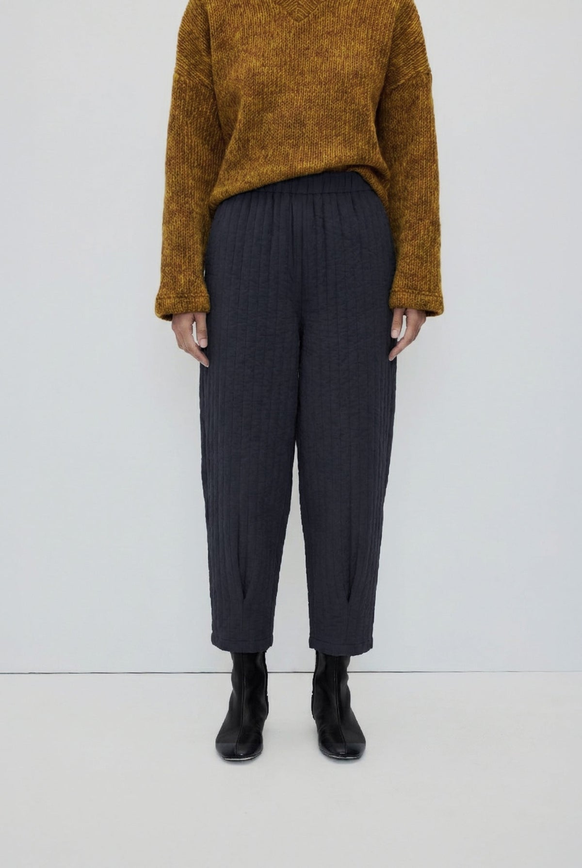 A model is shown from the chest down wearing dark charcoal gray pleated cropped trousers, a textured mustard yellow sweater, and black ankle boots against a plain white studio background.