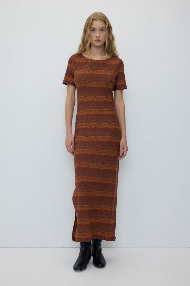 A woman modeling a short-sleeve knit maxi dress with horizontal rust-brown and orange stripes, paired with black ankle boots, against a plain white studio background.