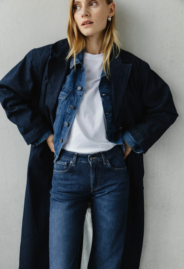 Person wearing a long dark coat over a denim jacket and jeans against a plain background