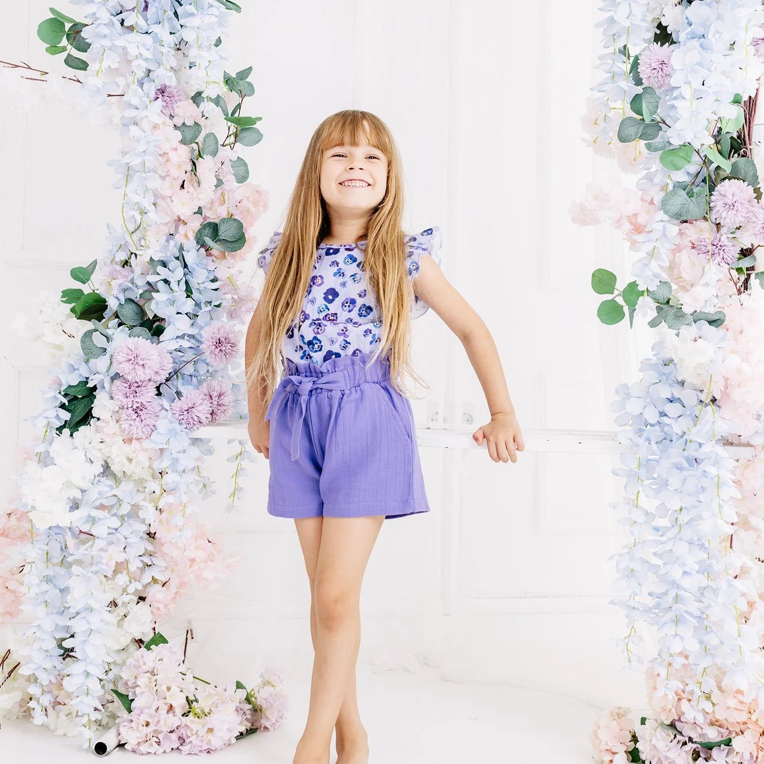 Young girl in a floral top and purple shorts standing between floral decorations.