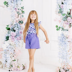 Young girl in a floral top and purple shorts standing between floral decorations.