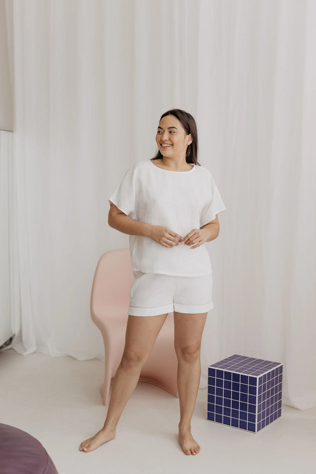 Woman in white shirt and shorts standing in a room with a pink chair and blue cube.