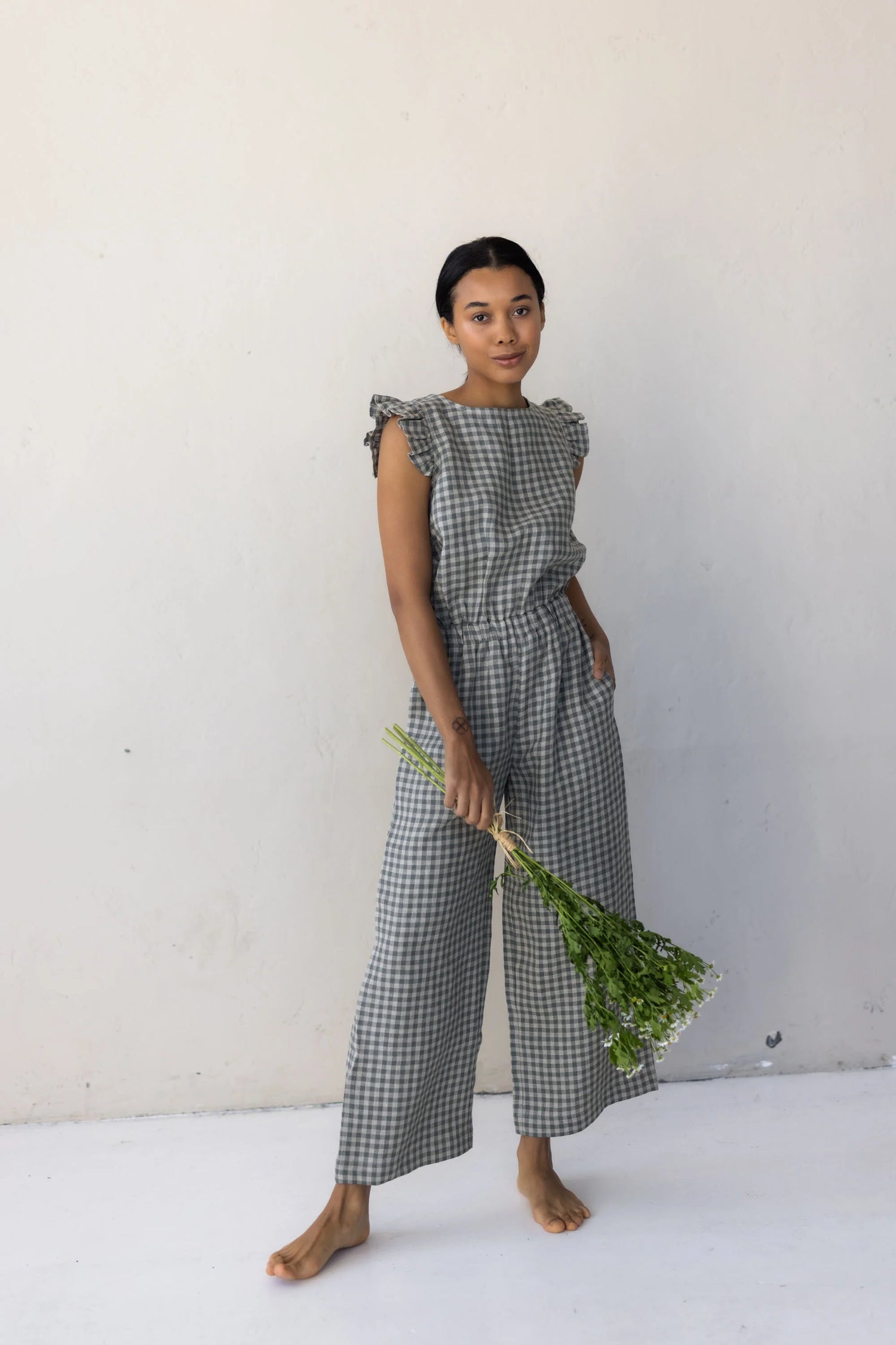 Woman wearing a checkered linen jumpsuit holding greenery against a plain background