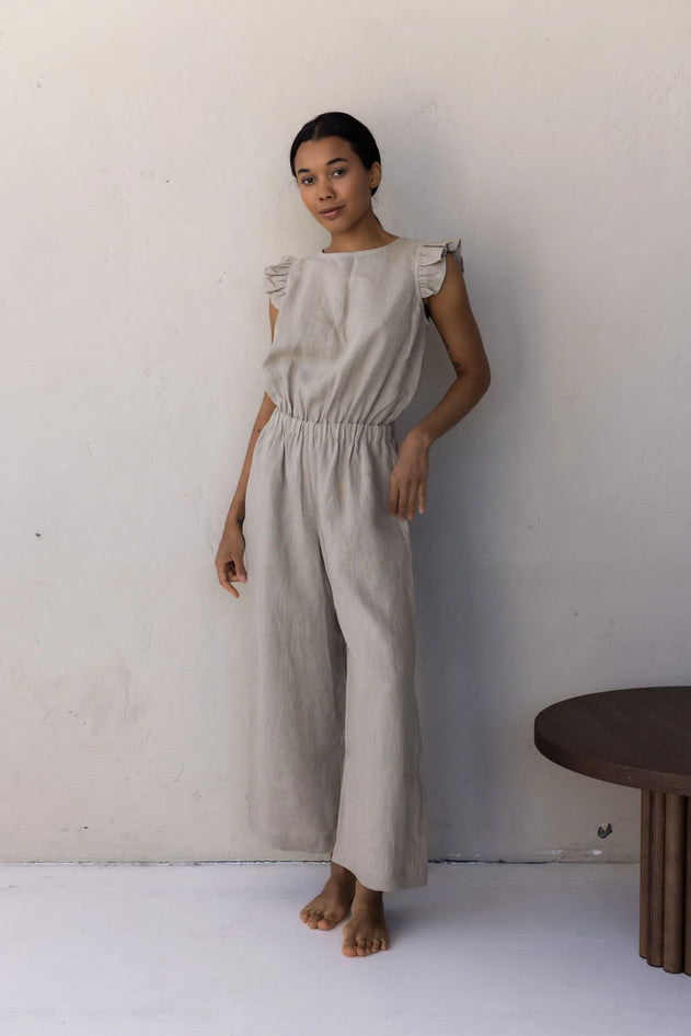 Woman wearing a beige jumpsuit standing against a white wall with a wooden table in the corner.