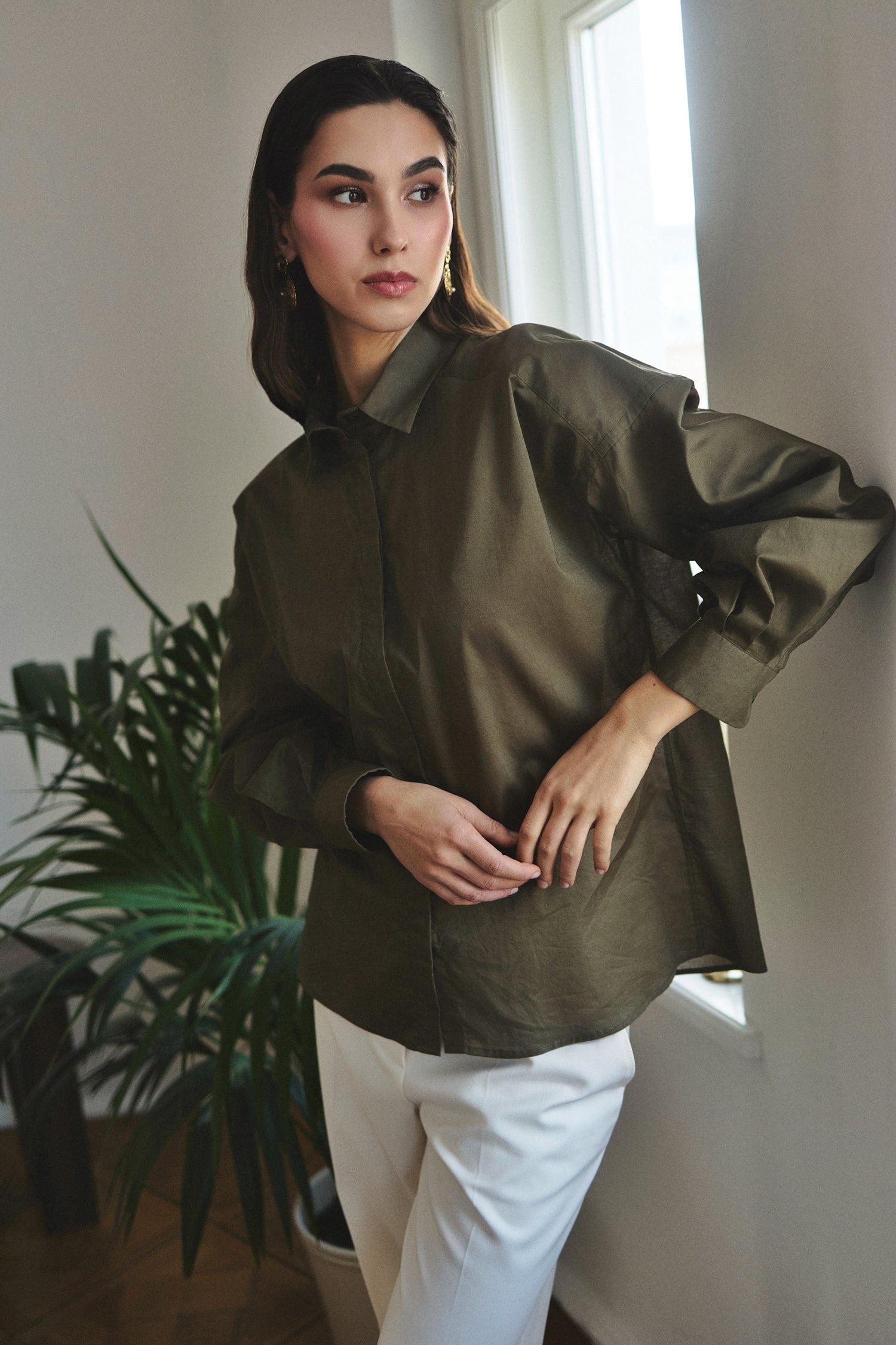 Woman wearing a green jacket and white pants standing indoors with a plant and window in the background.