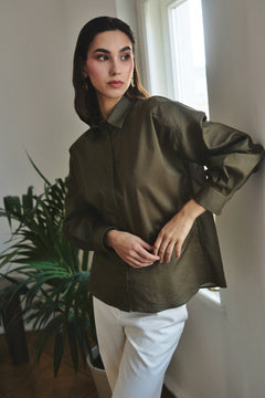 Woman wearing a green jacket and white pants standing indoors with a plant and window in the background.