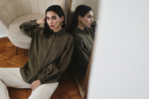 Woman sitting on a couch wearing a green shirt and white pants, reflected in a mirror.