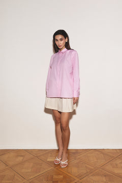 Woman wearing a light pink shirt and beige skirt standing against a white wall.