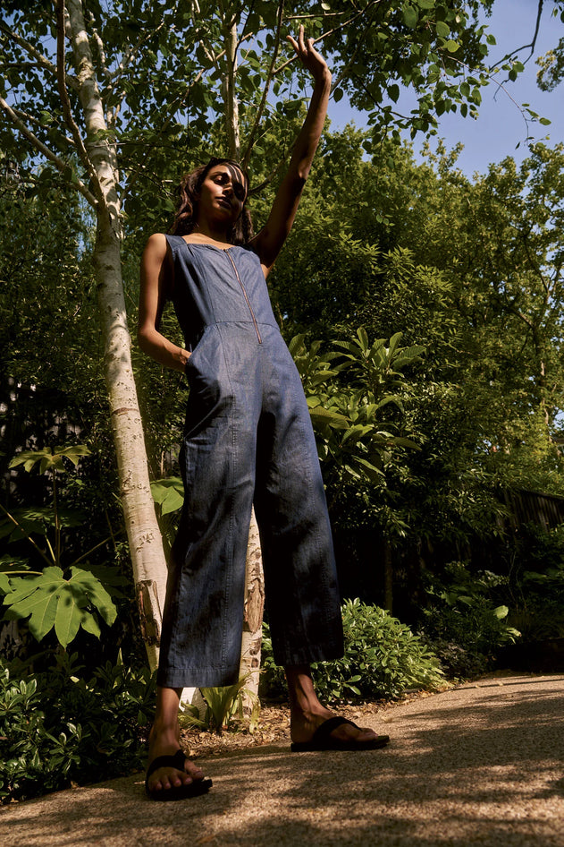 Woman in a denim jumpsuit standing in a forested area with trees and greenery around.