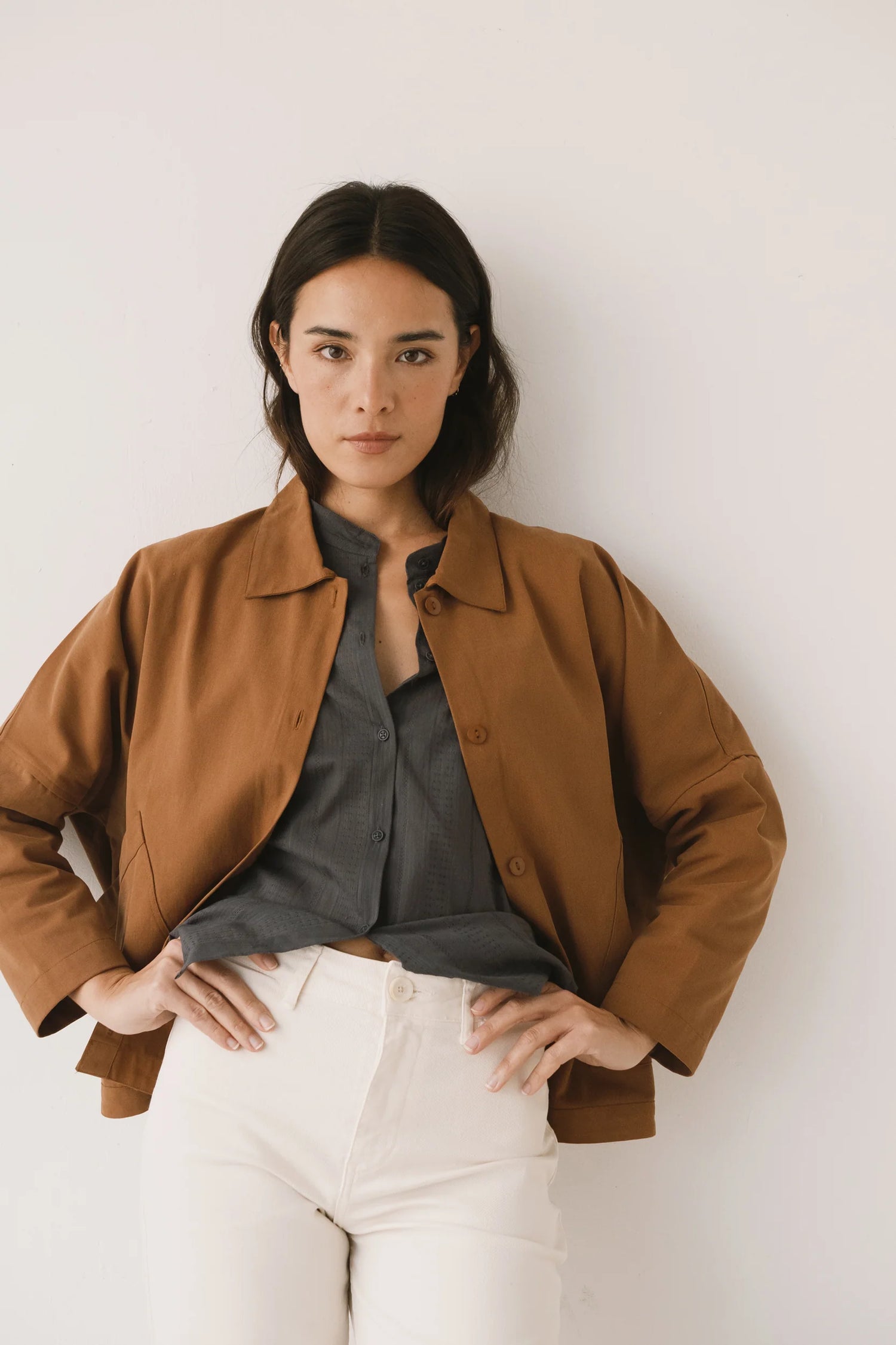 Woman wearing a brown jacket over a gray shirt with white pants against a plain background