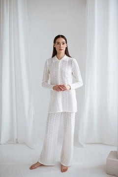 Woman wearing a white lace outfit standing against a white curtain.