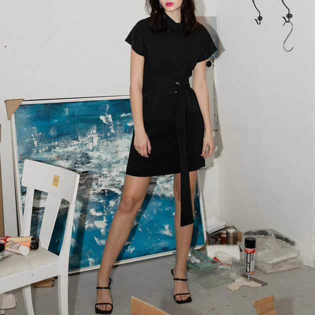 Person wearing a black dress standing in an art studio with a blue abstract painting in the background.