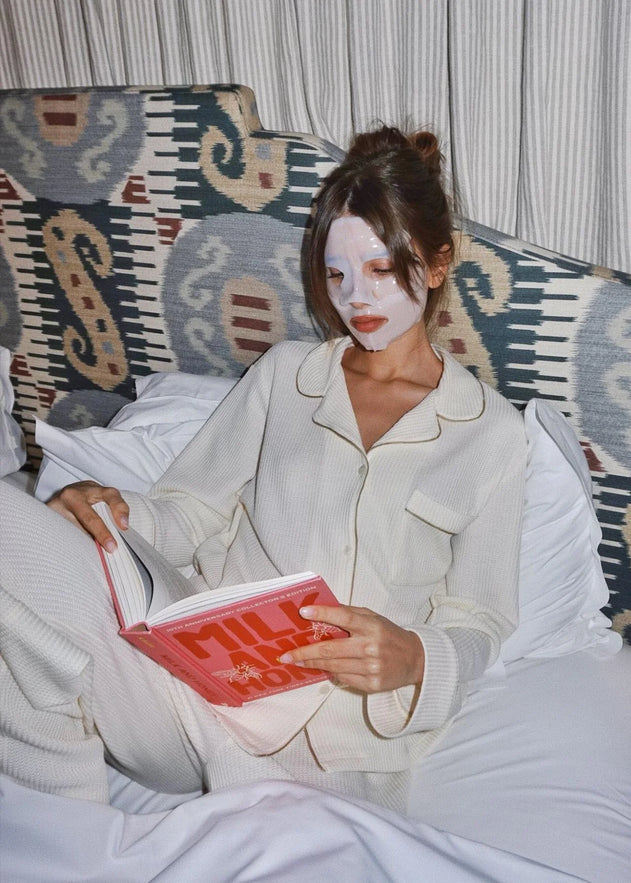 Person in pajamas with a face mask reading a book on a patterned couch.