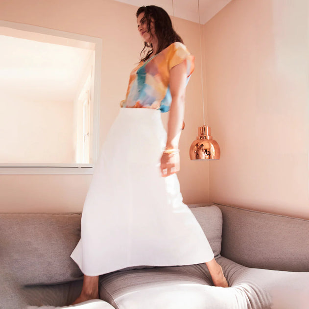 Woman in a colorful top and white skirt standing in a room with a lamp and window.