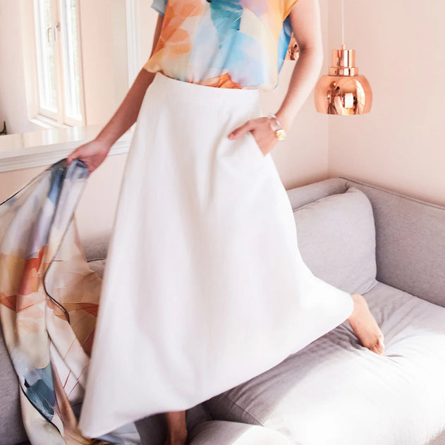 Person wearing a colorful top and white skirt in a room with a gray sofa and window.