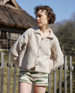 Woman wearing a beige knitted cardigan and patterned shorts standing outdoors with a wooden fence and thatched-roof building in the background.