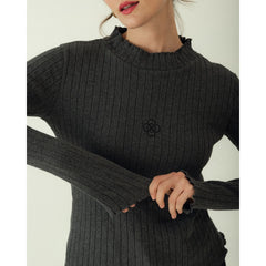 A close-up of a woman wearing a dark charcoal gray ribbed knit sweater with a small black embroidered floral logo on the chest, set against a plain light background.