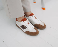 White low-top sneakers with brown suede panels and orange accents, shown on a person wearing light beige pants on a white floor.