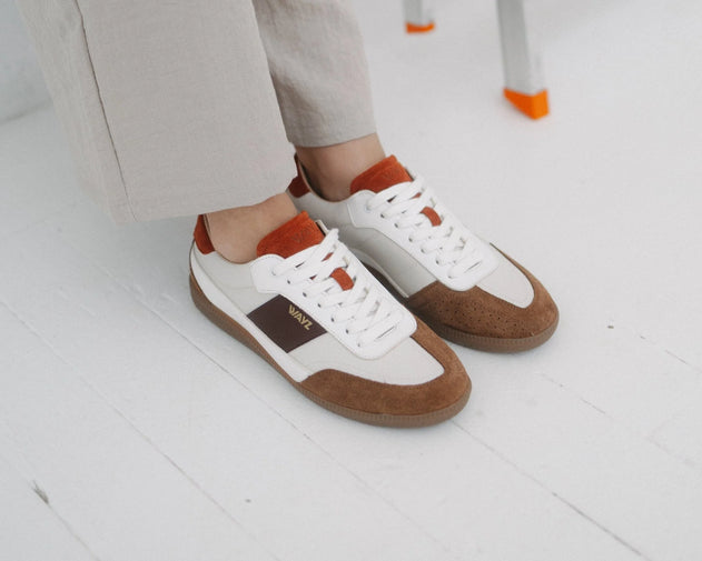White low-top sneakers with brown suede panels and orange accents, shown on a person wearing light beige pants on a white floor.