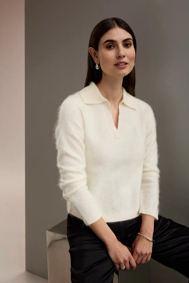 A woman wearing a fuzzy off-white long-sleeve polo-neck sweater and black trousers, seated against a neutral gray background.