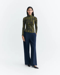 A woman stands against a plain light gray studio background wearing a fitted mock-neck knit top with an olive green and black plaid pattern, dark navy wide-leg trousers, and black pointed flats.