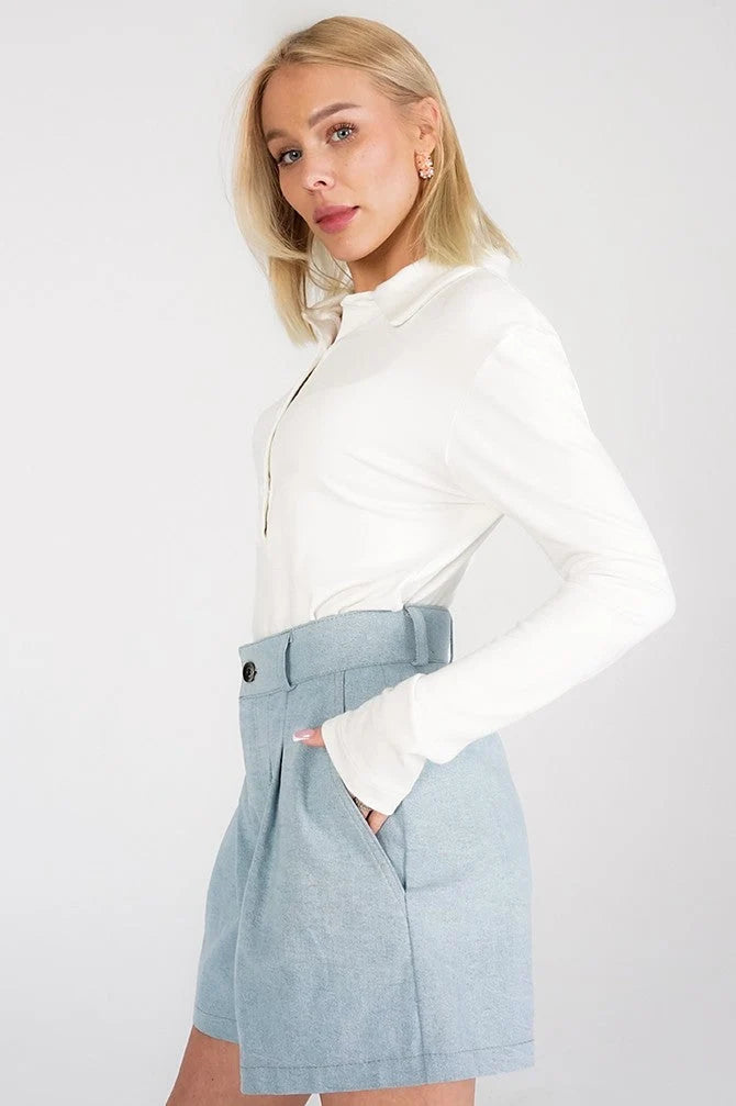 A blonde model wearing a white long-sleeve collared top and a light blue high-waisted pleated mini skirt with a matching belt, posed against a light gray studio background.