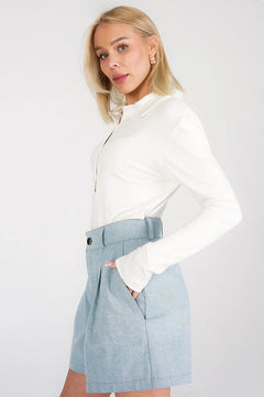 A blonde model wearing a white long-sleeve collared top and a light blue high-waisted pleated mini skirt with a matching belt, posed against a light gray studio background.