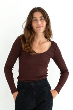A woman with long brown hair wearing a dark brown scoop-neck long-sleeve top and dark corduroy pants, standing against a plain white background.