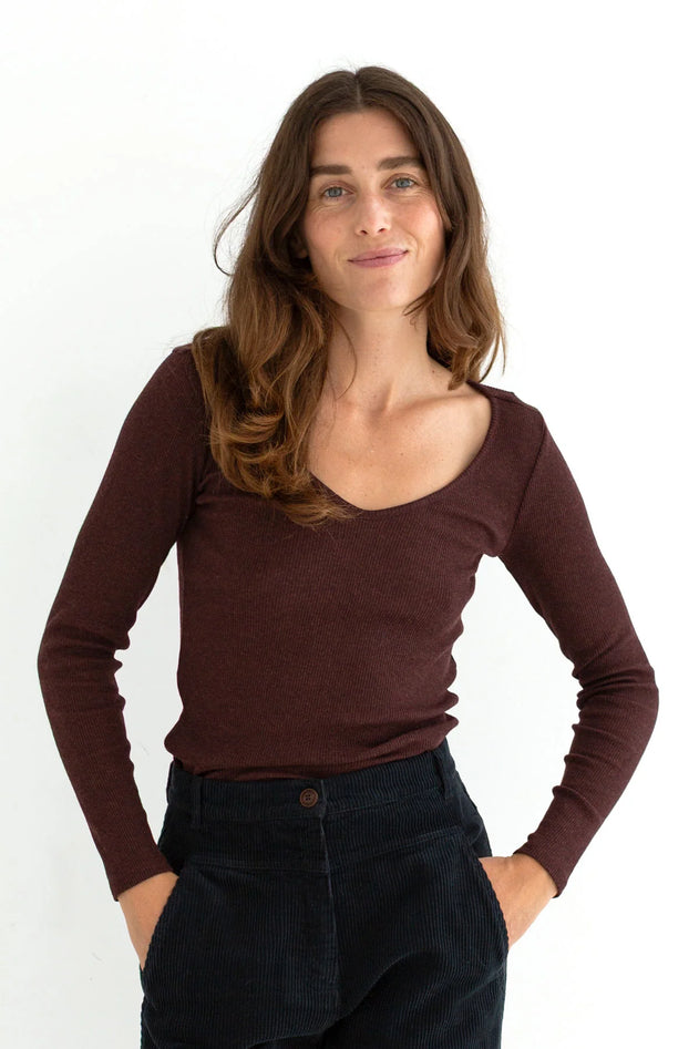 A woman with long brown hair wearing a dark brown scoop-neck long-sleeve top and dark corduroy pants, standing against a plain white background.