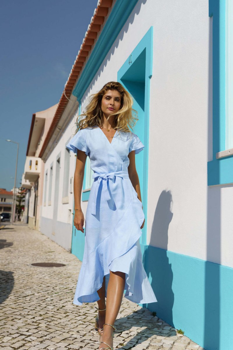 A blonde woman walks on a cobblestone street beside a white building with blue trim, wearing the Nakoa Annika Dress Sky Blue with an adjustable waistline and sandals on a sunny day.