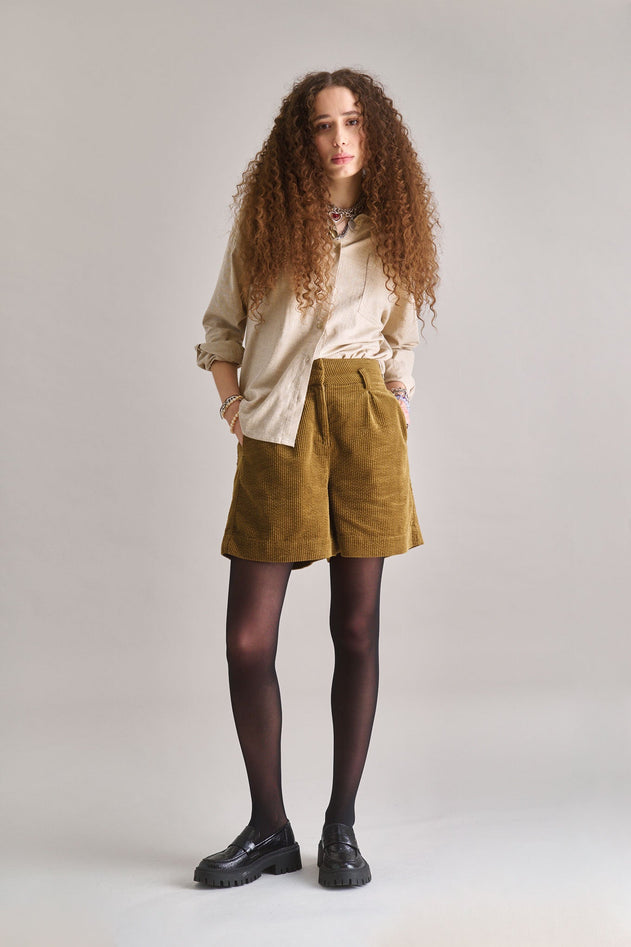Woman wearing a beige shirt, green shorts, and black shoes on a plain background