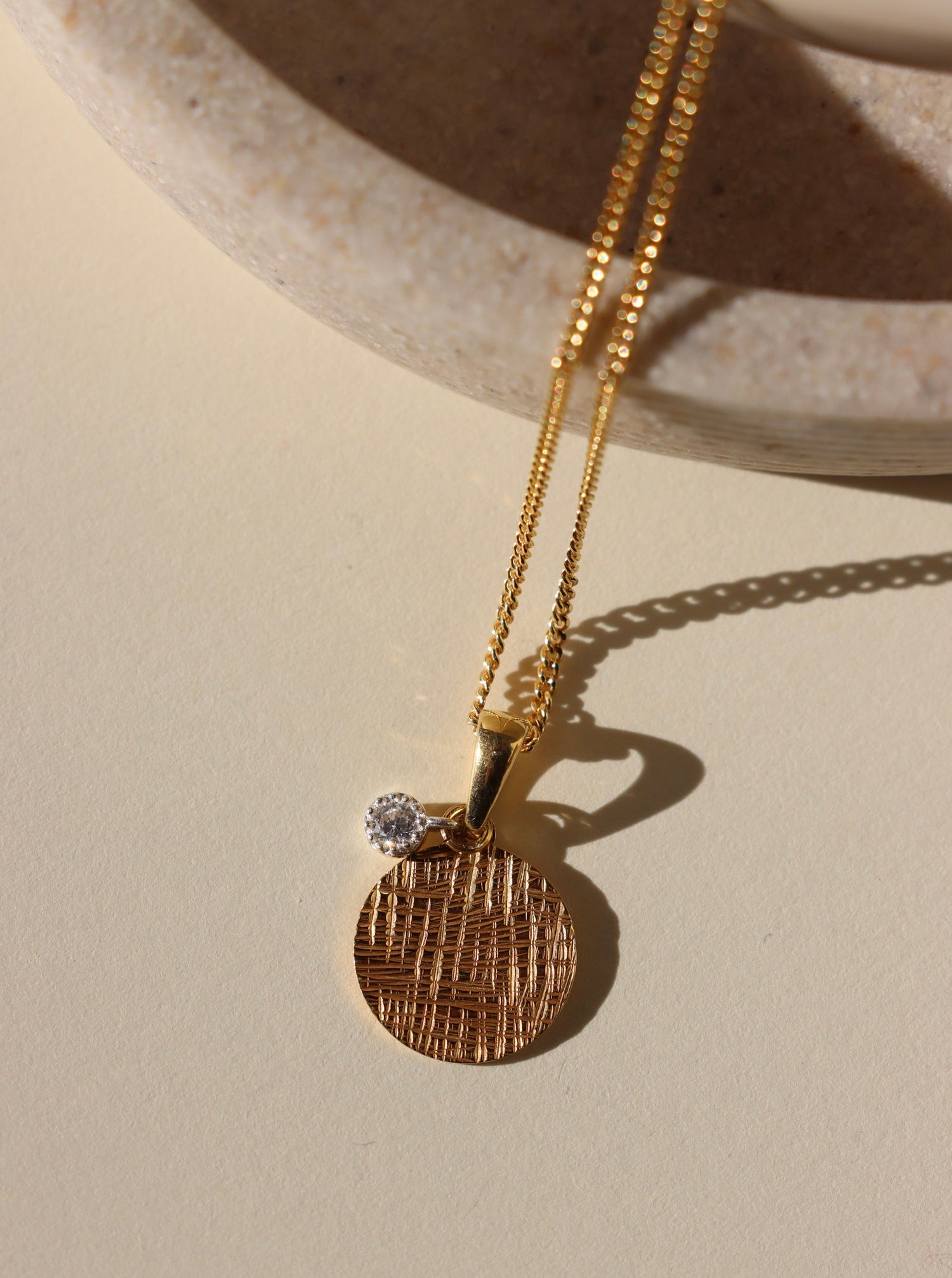 Gold necklace with a textured pendant and diamond on a beige surface