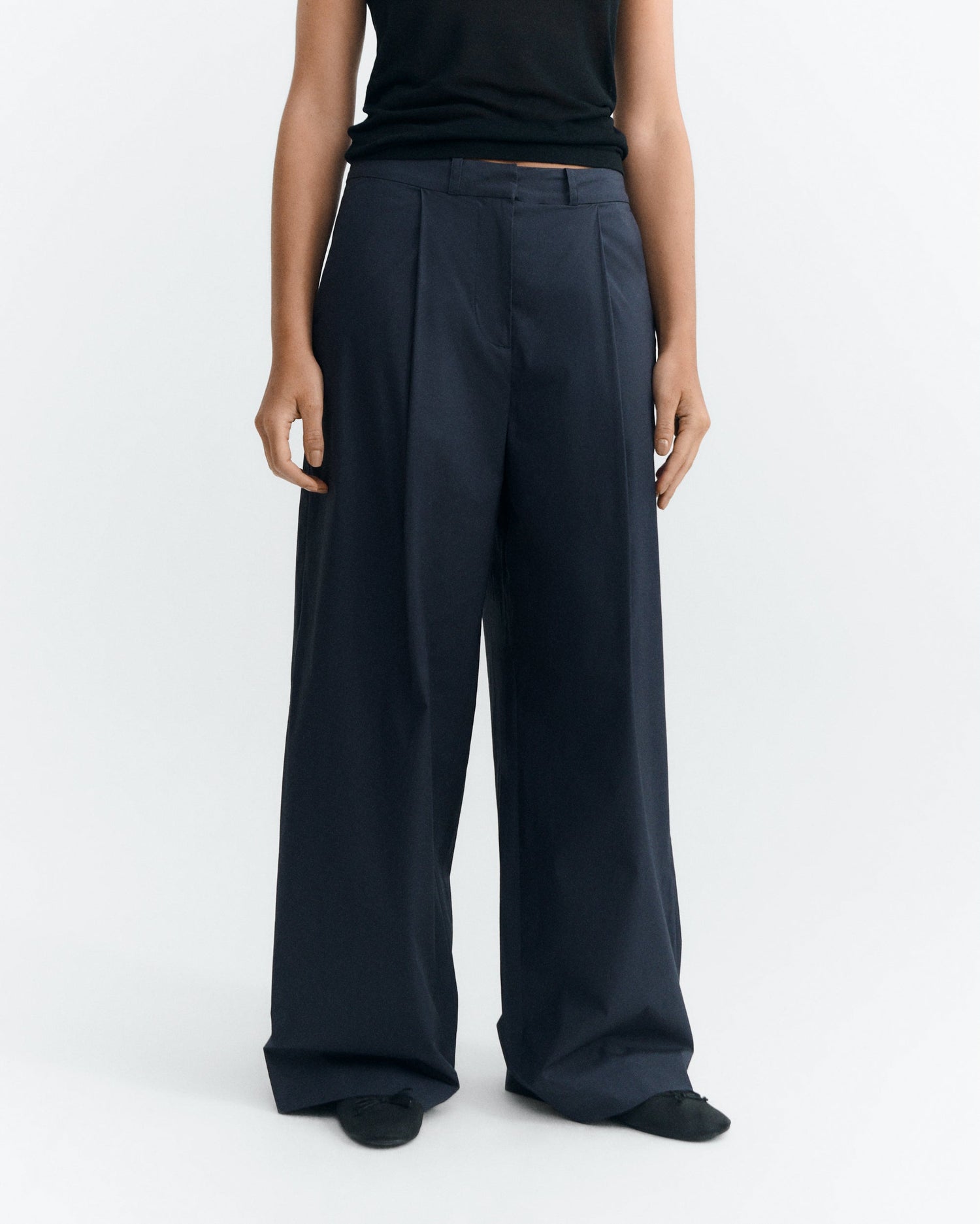 A model wearing a pair of dark navy wide-leg pleated trousers, paired with a black top and black shoes, photographed against a clean white studio background.