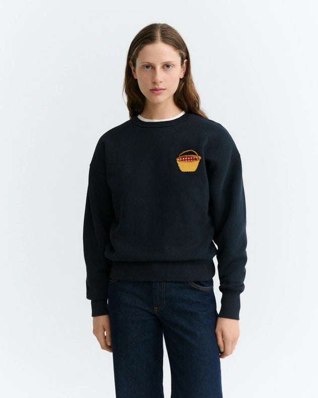A woman modeling a dark navy crewneck sweatshirt with a small embroidered yellow and red basket graphic on the left chest, paired with dark blue jeans, against a plain light gray background.