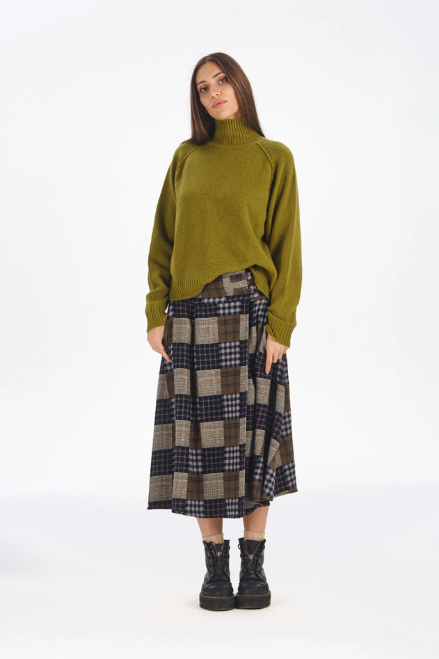 A woman stands against a plain white studio background wearing an olive green knit turtleneck sweater, a brown and navy plaid midi skirt, and black lace-up ankle boots.