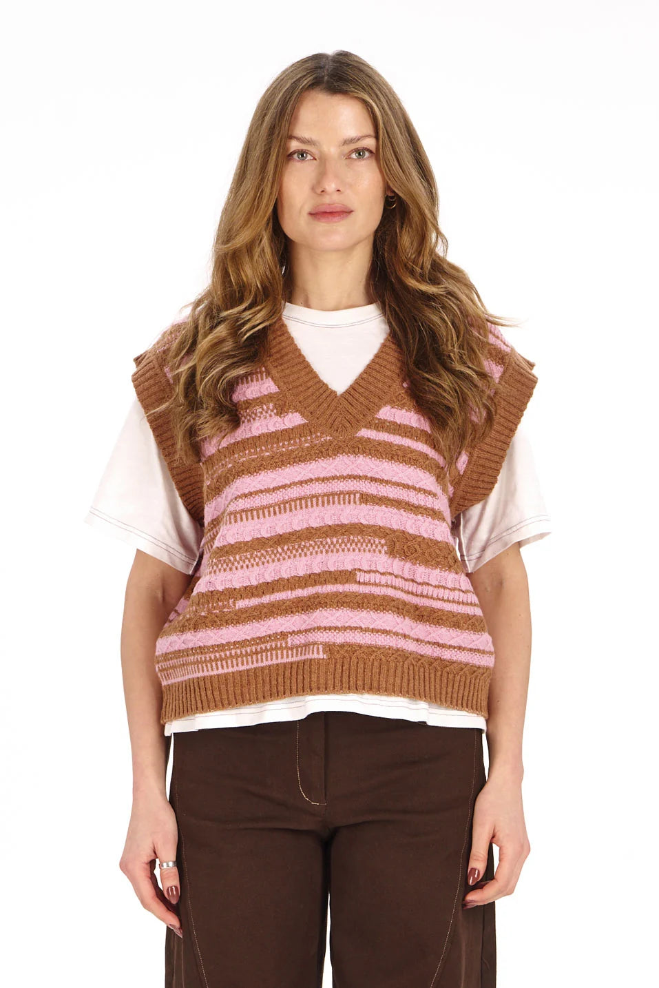 A woman modeling a brown and pink striped knit sweater vest over a white t-shirt and dark brown pants, shot against a solid white background.