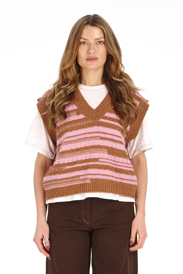 A woman modeling a brown and pink striped knit sweater vest over a white t-shirt and dark brown pants, shot against a solid white background.