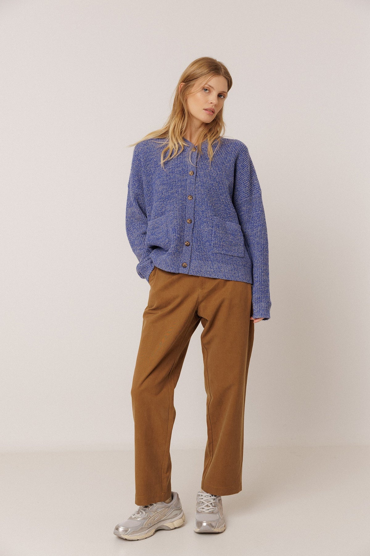 A woman modeling a blue textured button-up cardigan sweater, brown wide-leg trousers, and silver sneakers against a plain light gray studio background.