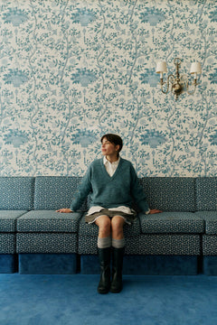 A person sits on a patterned blue sofa against blue floral wallpaper, wearing a teal sweater over a white collared shirt, a short skirt, and tall black boots.
